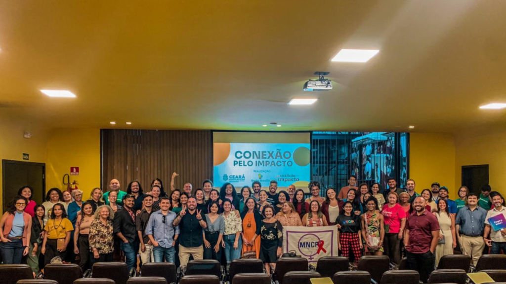 Foto de um grupo de pessoas reunidas em um auditório amarelo. No fundo, um telão exibe o título "Conexão pelo Impacto", acompanhado de logotipos de realizadores, incluindo o Social Brasilis e Coalizão pelo Impacto.